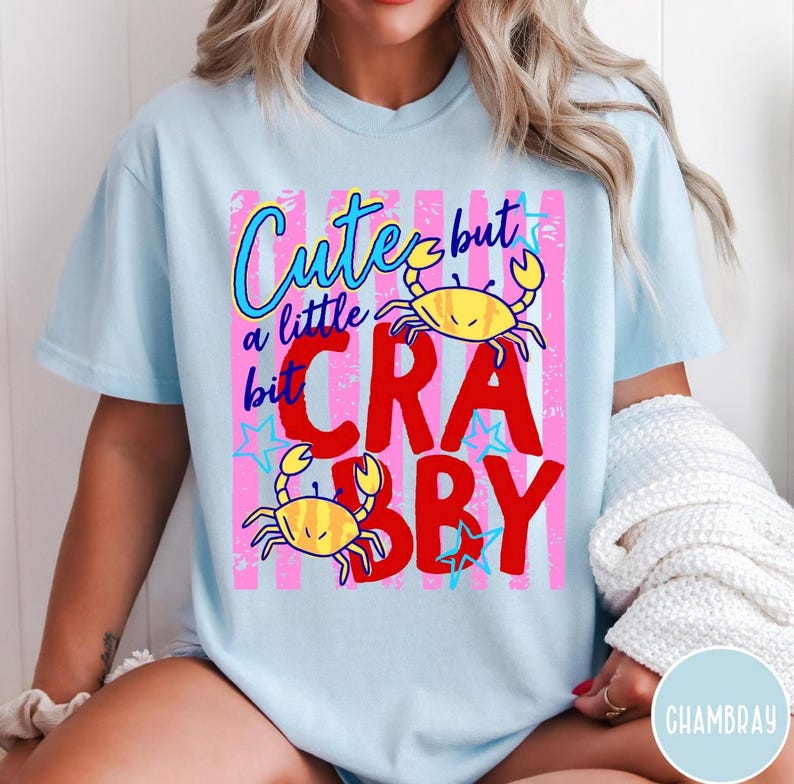 May include: A light blue t-shirt featuring a graphic design with pink vertical stripes, two yellow crabs, and blue stars. The text reads "Cute but a little bit CRABBY" in a mix of blue cursive and bold red block lettering across the center.