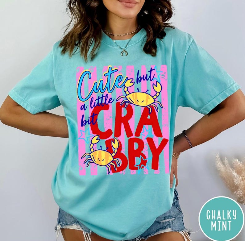 May include: A light turquoise short-sleeve t-shirt featuring a graphic print with the text "Cute but a little bit CRABBY" in blue and red. The design includes two yellow crabs and pink vertical stripes with blue star accents on the front.