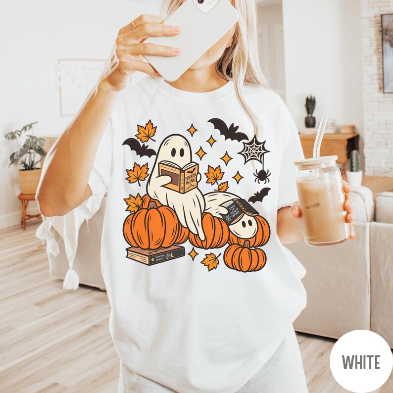 K&ouml;nnte beinhalten: Ein wei&szlig;es T-Shirt mit einem Grafikdruck von zwei Geistern, die B&uuml;cher lesen und zwischen orangefarbenen K&uuml;rbissen sitzen. Das Design enth&auml;lt orangefarbene Ahornbl&auml;tter, schwarze Flederm&auml;use, ein Spinnennetz und eine Spinne.