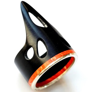 May include: A unique black ring with a pointed top and cut-out design. The ring features a band of orange and white material, set between silver edges. The ring's design is modern and eye-catching, suitable for a bold fashion statement.