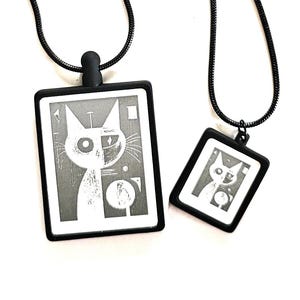 Könnte beinhalten: Zwei schwarze Katzenanhänger-Halsketten. Jeder Anhänger hat einen schwarzen Rahmen und ein Katzendesign in Weiß und Grau. Das Katzendesign ist abstrakt und geometrisch. Die Halsketten haben schwarze Ketten.