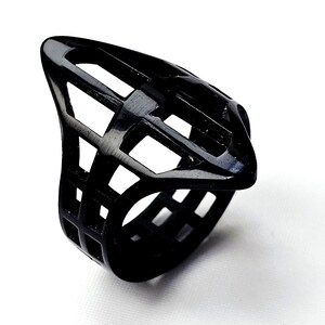May include: A black, geometric ring with a cage-like design. The ring features a series of rectangular cutouts, creating an open, airy appearance. The ring's shape is elongated and pointed, with a smooth, glossy finish.