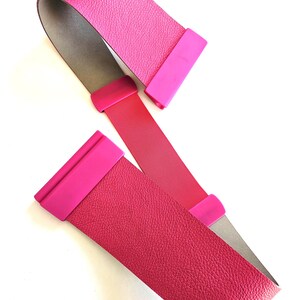 Könnte beinhalten: Ein leuchtend pinkfarbener Gürtel mit strukturierter Oberfläche. Der Gürtel hat einen roten Riemen und ist mit rechteckigen, pinkfarbenen Verschlüssen versehen. Das Design ist schlicht und modern.