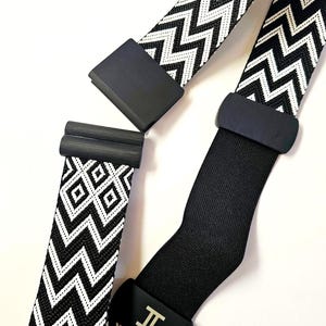 Puede incluir: Primer plano de tirantes estampados en blanco y negro con herrajes negros. Los tirantes presentan un patrón de zigzag y diamante. Un tirante tiene un símbolo metálico en el herraje negro.