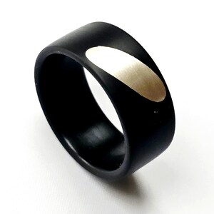 May include: A black ring featuring a silver oval inlay. The ring has a smooth, matte finish and a wide band. The silver inlay is positioned on the top of the ring.
