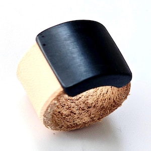 May include: A handcrafted leather cuff bracelet. The bracelet features a natural tan leather band with a rough, textured edge. A curved, black, rectangular piece is attached to the top of the band.
