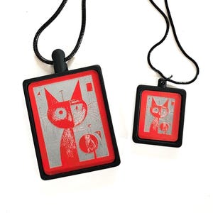 May include: Two pendant necklaces featuring a cat design. The pendants have a red border and a black frame. The cat design is red and silver. The necklaces have black cords.