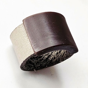 May include: A wide cuff bracelet with a two-tone design. The top section is a dark brown, curved shape, while the band is a textured, off-white color. The interior edge has a rough, dark texture. The bracelet is a bold accessory.