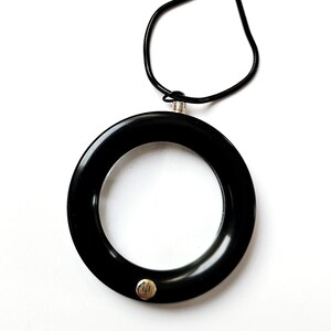 May include: A black circular pendant necklace with a silver-colored clasp and a thin black cord. The pendant has a small silver-colored stud with the letter "H" on it. The necklace is set against a white background.