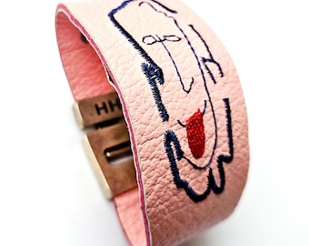 Op de afbeelding: Een lichtroze leren armband met een grillig geborduurd gezicht. Het gezicht heeft blauwe stiksels voor de contouren en details, met een rode tong. De armband heeft een metalen sluiting en de initialen "HH" zijn zichtbaar. Ongeveer 20 cm lang.