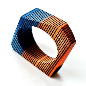 Könnte beinhalten: Geometrischer Holzring. Der Ring ist in blaue und orangefarbene Abschnitte mit schwarzen Linien unterteilt. Das Design ist modern und künstlerisch. Der Durchmesser beträgt ca. 2,5 cm.