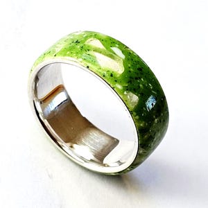 Puede incluir: Un anillo de banda ancha con interior plateado y exterior moteado verde y blanco. El anillo tiene un acabado liso y pulido y un diseño moderno. El anillo se muestra sobre un fondo blanco.