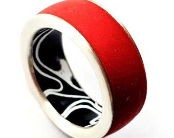 Puede incluir: Un anillo rojo con una banda plateada y un interior con estampado en blanco y negro. El anillo tiene un acabado liso y mate y un diseño moderno. El anillo es una joya.