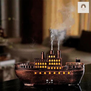 May include: A bronze-toned ship-shaped essential oil diffuser, featuring illuminated windows and smokestacks. The diffuser releases a stream of vapor, creating a relaxing atmosphere. The design is reminiscent of a vintage steamship, adding a nautical touch to any room.