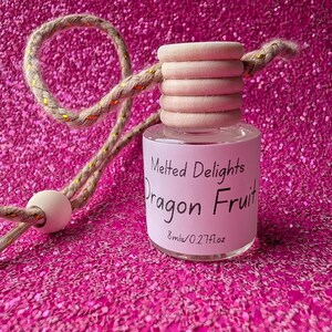May include: A small glass bottle with a wooden cap and a decorative cord. The bottle has a pink label with the text "Melted Delights Dragon Fruit" and measures 8 ml / 0.27 fl oz. The background is a sparkly pink surface.