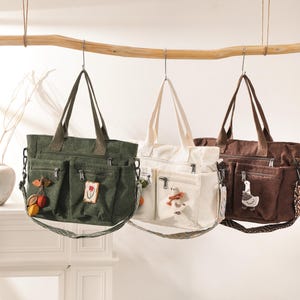 May include: Three corduroy tote bags in olive green, cream, and brown hang from a wooden rod. Each bag has multiple pockets, a shoulder strap, and decorative patches. The bags are displayed against a neutral background.