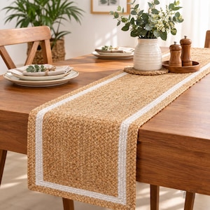May include: A rectangular table runner made of braided natural tan jute fibre with a white fabric border. The runner is placed on a wooden table and features a textured, woven surface design that runs along the centre of the dining furniture.