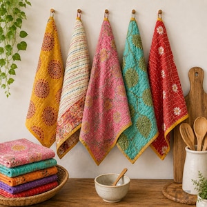May include: Five quilted kitchen towels hang from hooks, featuring patterns in yellow, white, pink, teal, and red. A stack of folded towels in pink, teal, orange, purple, and red sits in a woven basket next to a small ceramic bowl and wooden spoons.