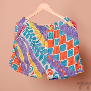 Könnte beinhalten: Bunte Patchwork-Shorts mit einer lebendigen Mischung aus Mustern. Die Shorts zeigen eine Kombination aus orangefarbenen, lila, gelben und blauen geometrischen und floralen Designs. Die Shorts hängen an einem Holzbügel.