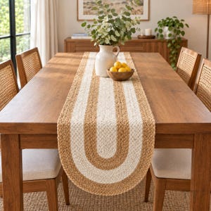 Può includere: Un runner da tavola rettangolare in juta intrecciata con una striscia centrale bianca a forma di U. Il runner è posizionato su un tavolo da pranzo in legno con quattro sedie, un vaso in ceramica bianca con fogliame e una ciotola di limoni.