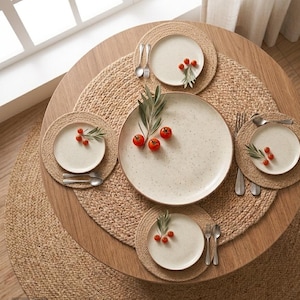 Op de afbeelding: Een ronde houten eettafel gedekt met een groot centraal bord en vier kleinere borden, elk geplaatst op een geweven jute placemat. De gebroken witte gespikkelde keramische borden zijn versierd met kleine rode cherrytomaatjes en olijftakjes.