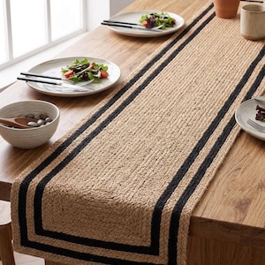Pode incluir: Um caminho de mesa retangular feito de fibras de juta natural entrançadas em tom bege, com duas faixas pretas paralelas que percorrem todo o comprimento. O caminho de mesa está colocado sobre uma mesa de madeira com pratos e taças.
