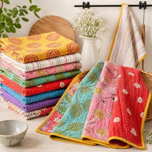 May include: A stack of folded cotton quilts featuring various patterns including yellow circles, pink florals, green leaves, and red daisies. Each quilt is finished with visible running stitch quilting and a solid yellow fabric border.