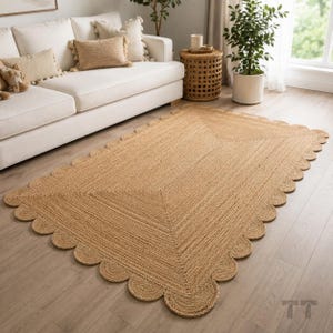 Op de afbeelding: Een rechthoekig vloerkleed gemaakt van natuurlijke, lichtbruine jutvezels, met een geweven textuur en een decoratieve geschulpte rand. Het kleed ligt plat op een lichte houten vloer en vertoont een consistent gevlochten patroon.