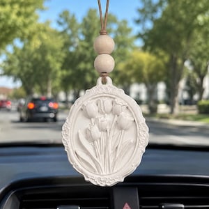 May include: A white, oval-shaped hanging car ornament featuring a relief design of a bouquet of tulips. The ornament is suspended by a brown leather cord accented with two round, light-colored wooden beads, hanging from a rearview mirror.