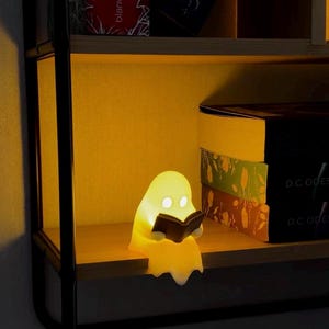 May include: A small, glowing, yellow-tinted ghost-shaped lamp sits on a wooden shelf. The ghost figure has two circular white eyes and is holding a small, dark, open book in its hands. The lamp emits a warm, soft light that illuminates the surrounding area.