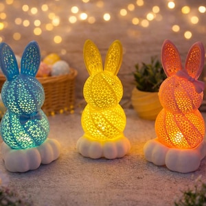 Könnte beinhalten: Drei beleuchtete Osterhasen-Dekorationen in Blau, Gelb und Orange. Jeder Hase hat ein gemustertes Design und sitzt auf einer weißen Wolkenbasis. Ostereier und ein Korb sind im Hintergrund.