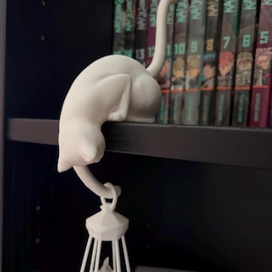 May include: A matte white figurine of a cat perched on a ledge with its tail hanging down. The cat's tail is curved to hold a small, white, cage-like lantern structure that hangs vertically beneath the shelf, featuring a pointed top and base.