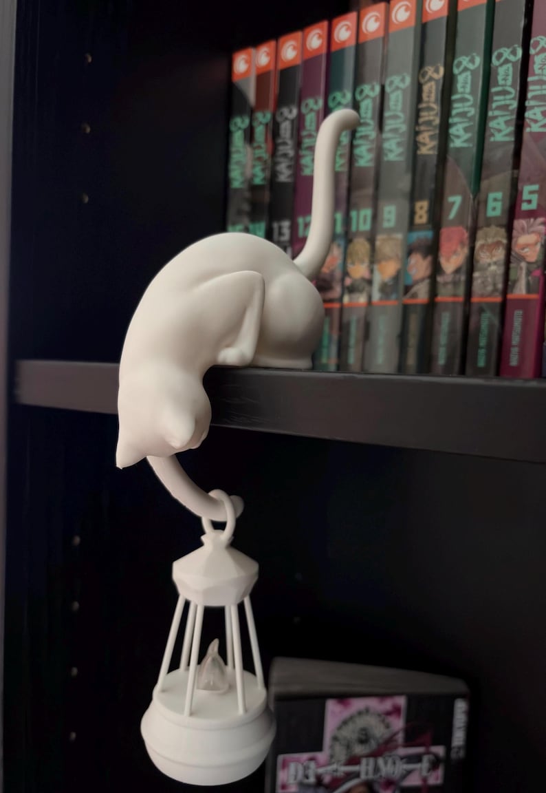 May include: A matte white figurine of a cat perched on a ledge with its tail hanging down. The cat's tail is curved to hold a small, white, cage-like lantern structure that hangs vertically beneath the shelf, featuring a pointed top and base.