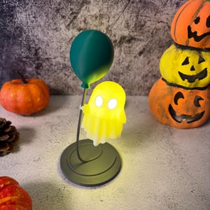 May include: A Halloween lamp with a glowing yellow ghost figure held by a dark green balloon. The lamp is on a circular base. Orange pumpkins are in the background, creating a festive atmosphere.