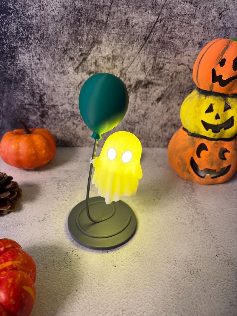 May include: A Halloween lamp with a glowing yellow ghost figure held by a dark green balloon. The lamp is on a circular base. Orange pumpkins are in the background, creating a festive atmosphere.