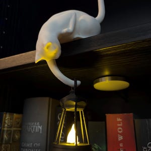 May include: A white cat figurine with a smooth matte finish, positioned on a shelf edge. The cat's tail hangs down, supporting a small black lantern that emits a warm yellow light from within, illuminating the cat's head and the lantern's interior.