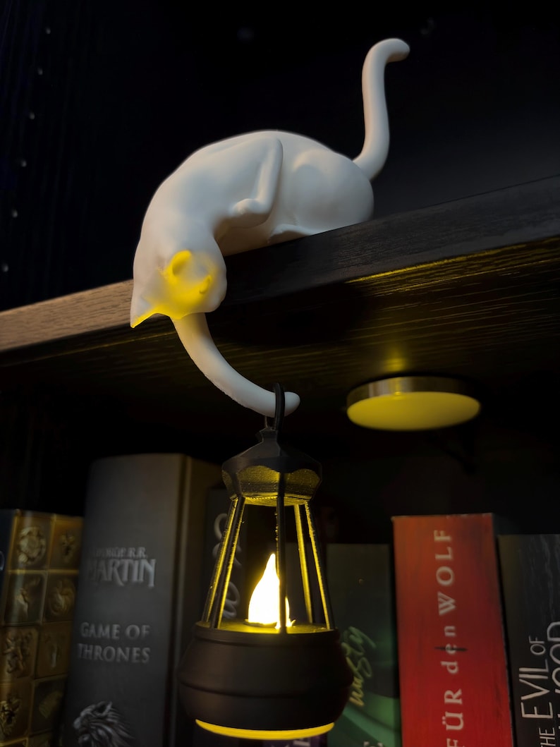 May include: A white cat figurine with a smooth matte finish, positioned on a shelf edge. The cat's tail hangs down, supporting a small black lantern that emits a warm yellow light from within, illuminating the cat's head and the lantern's interior.