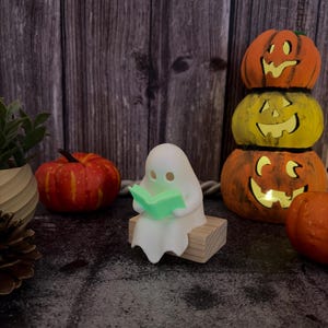 May include: A small white ghost figurine sitting on a rectangular wooden block, holding an open light green book. The ghost has two simple circular eye cutouts and is positioned among various orange and yellow decorative pumpkins.