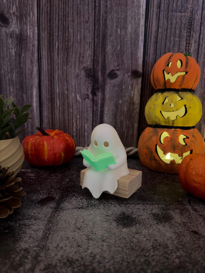 May include: A small white ghost figurine sitting on a rectangular wooden block, holding an open light green book. The ghost has two simple circular eye cutouts and is positioned among various orange and yellow decorative pumpkins.