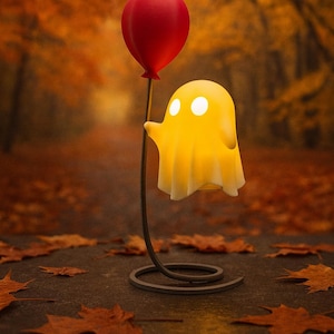 Könnte beinhalten: Eine skurrile Geisterlampe mit einem roten Ballon. Die gelbe Lampe hat zwei weiße, leuchtende Augen und wird von einem dünnen, gebogenen Metallständer gehalten. Die Lampe steht vor einem verschwommenen Herbsthintergrund mit fallenden Blättern.