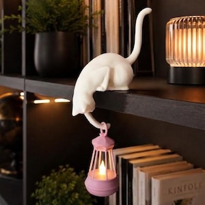 May include: A white cat figurine perched on the edge of a shelf, featuring a long tail that curves downward to hold a small, light pink lantern. The lantern contains a glowing, warm-toned LED light, creating a soft illumination effect.