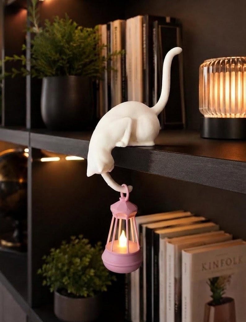 May include: A white cat figurine perched on the edge of a shelf, featuring a long tail that curves downward to hold a small, light pink lantern. The lantern contains a glowing, warm-toned LED light, creating a soft illumination effect.