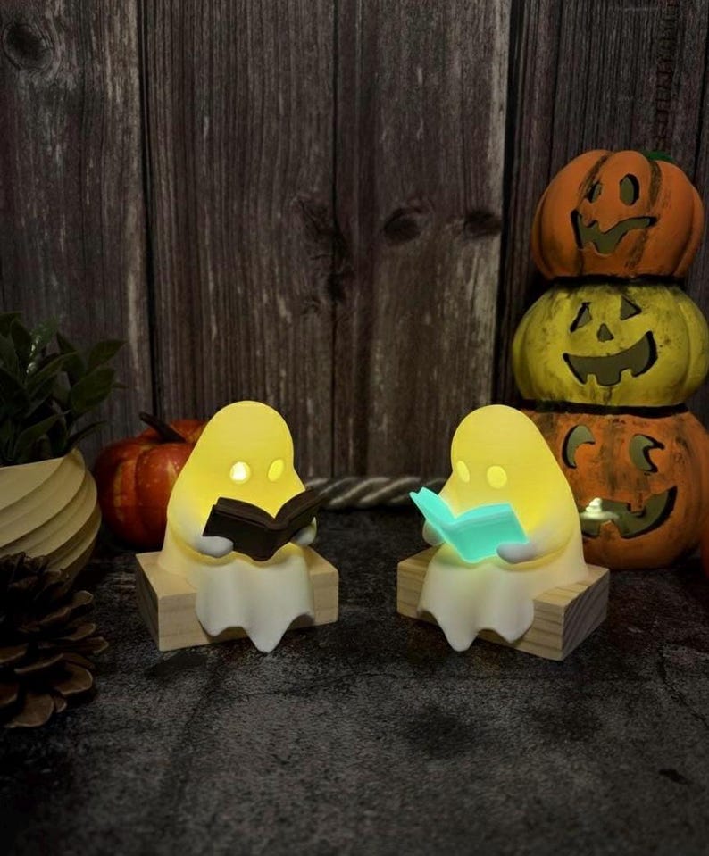 May include: Two small, glowing, off-white ghost figurines sitting on wooden blocks. Each ghost has illuminated eyes and holds an open book, one with a dark cover and the other with a bright teal cover, creating a soft, warm light effect.