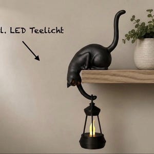 Könnte beinhalten: Eine schwarze Katzenfigur, die auf einer Regalbrettkante sitzt und mit ihrem Schwanz eine schwarze Laterne mit einem gelben LED-Teelicht hält. An der Wand befindet sich der Text "inkl. LED Teelicht" mit einem Pfeil, der auf die Figur zeigt.