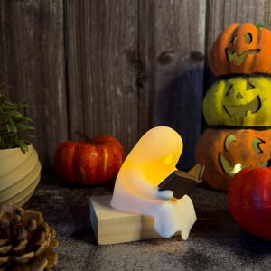 May include: A small, white, glowing ghost figurine sitting on a light-colored wooden block while holding and reading a miniature black book. The ghost emits a warm, soft yellow light from within its translucent, rounded body.
