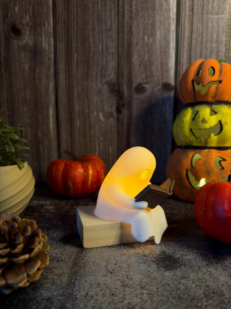 May include: A small, white, glowing ghost figurine sitting on a light-colored wooden block while holding and reading a miniature black book. The ghost emits a warm, soft yellow light from within its translucent, rounded body.