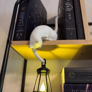 May include: A white cat figurine resting on a shelf, holding a black lantern with a glowing yellow light in its tail. The cat is positioned between two black books titled Dark and Very Bad, with a third book visible on the shelf below.