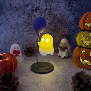 May include: A Halloween display with a glowing yellow ghost lamp holding a blue balloon. Other decorations include carved pumpkins, ghost figurines, and pine cones, all against a textured background. The lamp is on a black base.