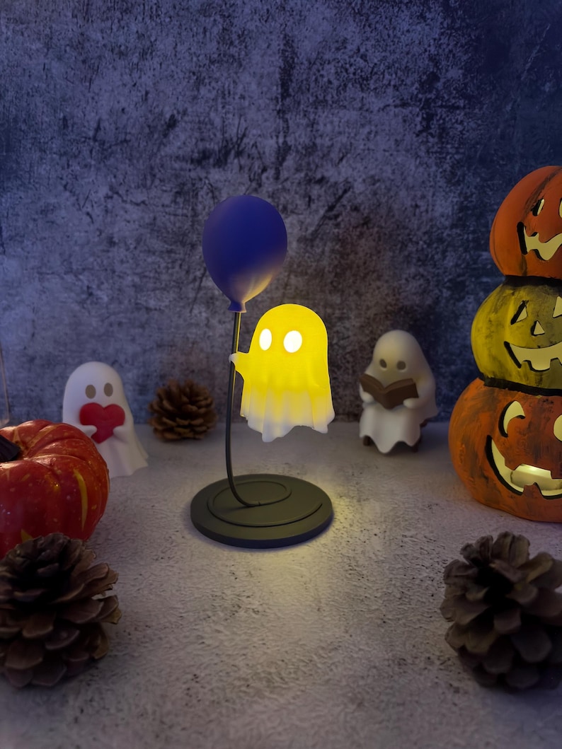 May include: A Halloween display with a glowing yellow ghost lamp holding a blue balloon. Other decorations include carved pumpkins, ghost figurines, and pine cones, all against a textured background. The lamp is on a black base.