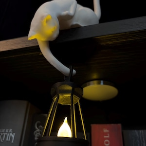 May include: A white cat figurine perched on a ledge, with its tail extending downward to hold a black lantern. The lantern features a glowing yellow light inside, casting a warm illumination on the cat's head and the surrounding area.